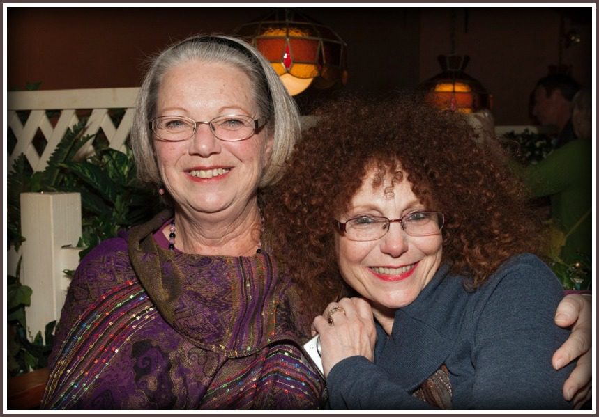 Two Individuals Closely Embracing Each Other Indoors, With One Wearing a Patterned Purple Shirt With Glitter Details and a Pearl Necklace, the Other in a Navy Blue Top.