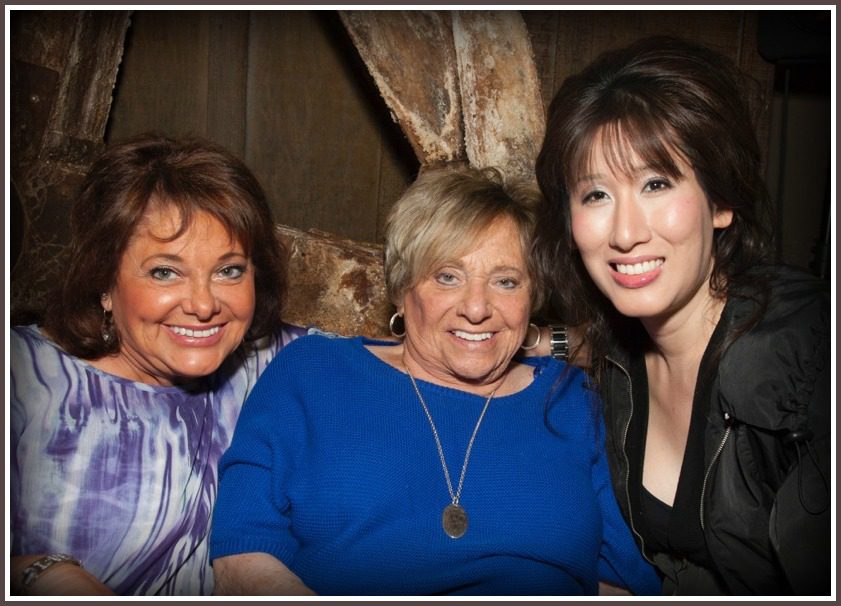 Three People Sitting Close Together in Front of a Rustic Background, With the Person in the Middle Wearing a Blue Sweater and a Silver Necklace and the Other Two Wearing Purple and Black Clothing.