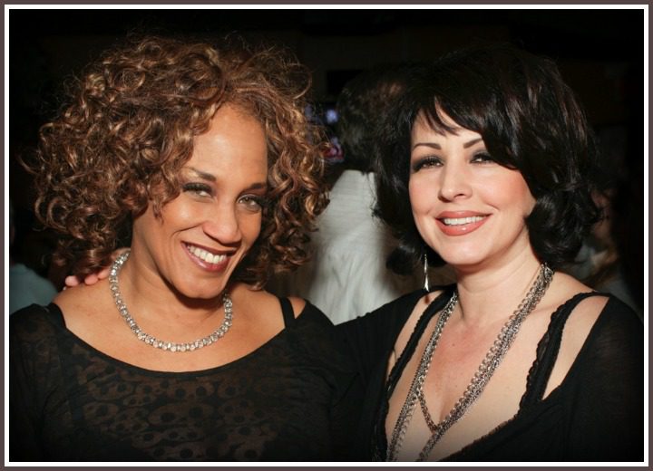 Two Women Standing Close Together, One With Curly Hair Wearing a Black Dress With a Sheer Patterned Overlay and a Silver Necklace, and the Other With Dark Hair in a Black Dress With Lacy Details and a Silver Chain Necklace.