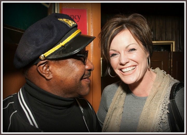 Two Individuals Standing Close to Each Other, With the One on the Left Wearing a Black Cap With Gold Trim and a Black Jacket With White Stripes on the Collar and Sleeve, and the One on the Right Wearing a Grey Top With a Creamy Lacy Detail and Carrying a Black Shoulder Bag.